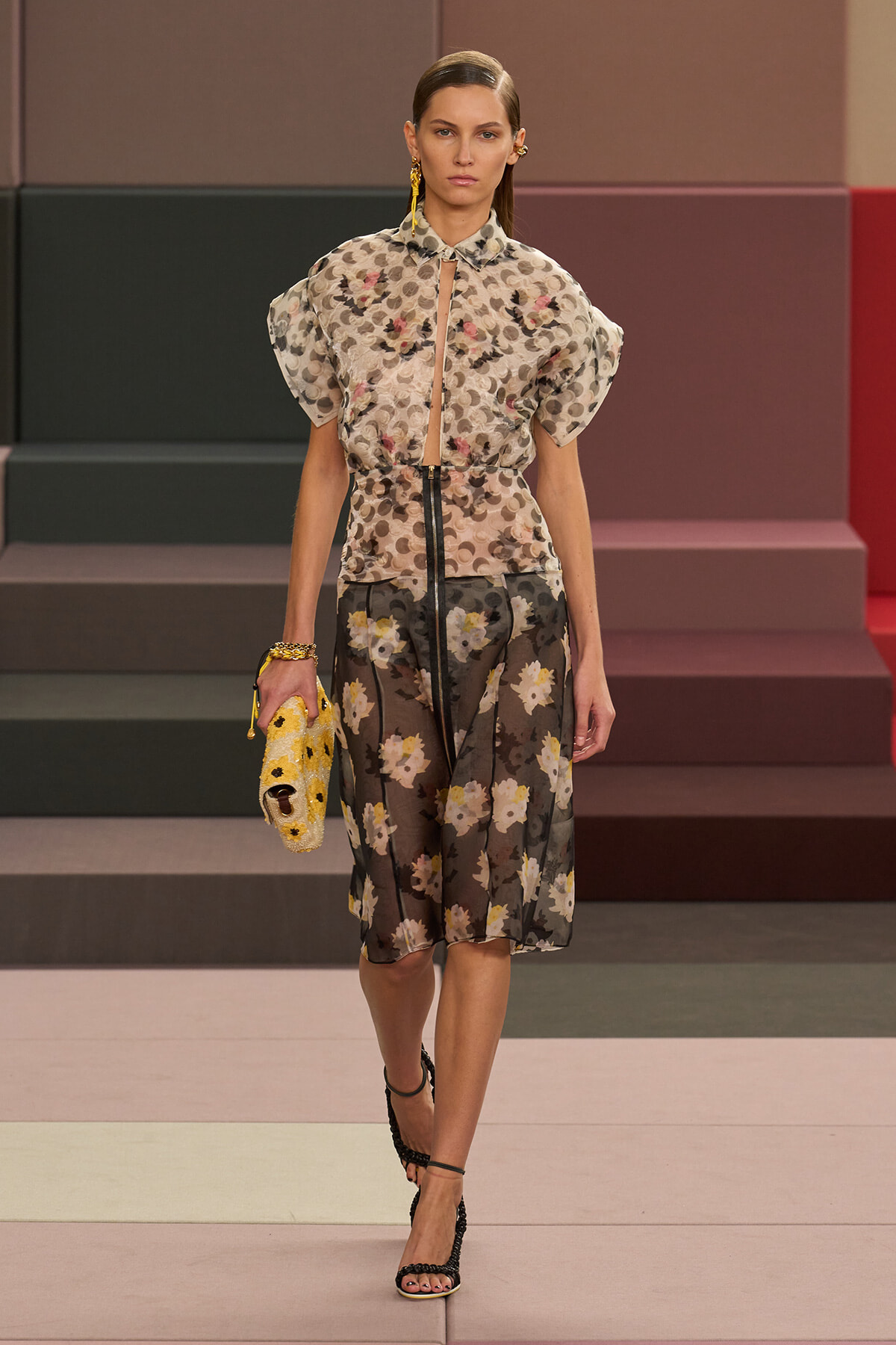 Runway model in a sheer floral blouse with puff sleeves and matching sheer floral skirt, holding a yellow embroidered clutch with black sandals.