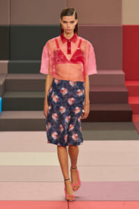 Model on a runway wearing a pink sheer top over a red bralette and a navy floral midi skirt, pink sandals visible under her feet.