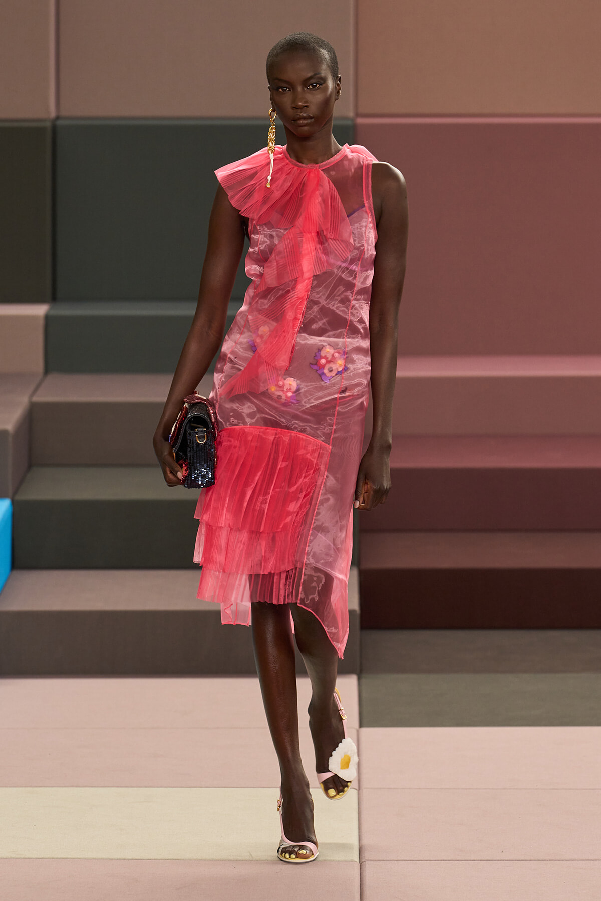 Model walking a runway in a pink sheer dress with layered ruffles and floral embroidery, carrying a small dark clutch and wearing pink heeled sandals with a white flower detail.