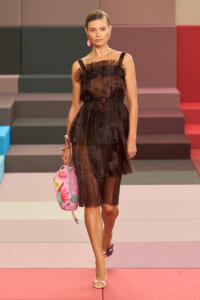 Model walking the runway in a sheer, pleated black dress with a pink floral bag.
