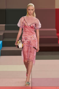 Model walking a pink sheer dress with floral embroidery on a color-blocked runway, carrying a small handbag.