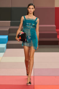 Model walking a fashion show in a teal sheer pleated dress, carrying a black floral clutch.