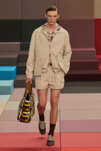 Male model walking a fashion show in a beige zip-up jacket and matching shorts, carrying a multi-colored checkered tote bag.