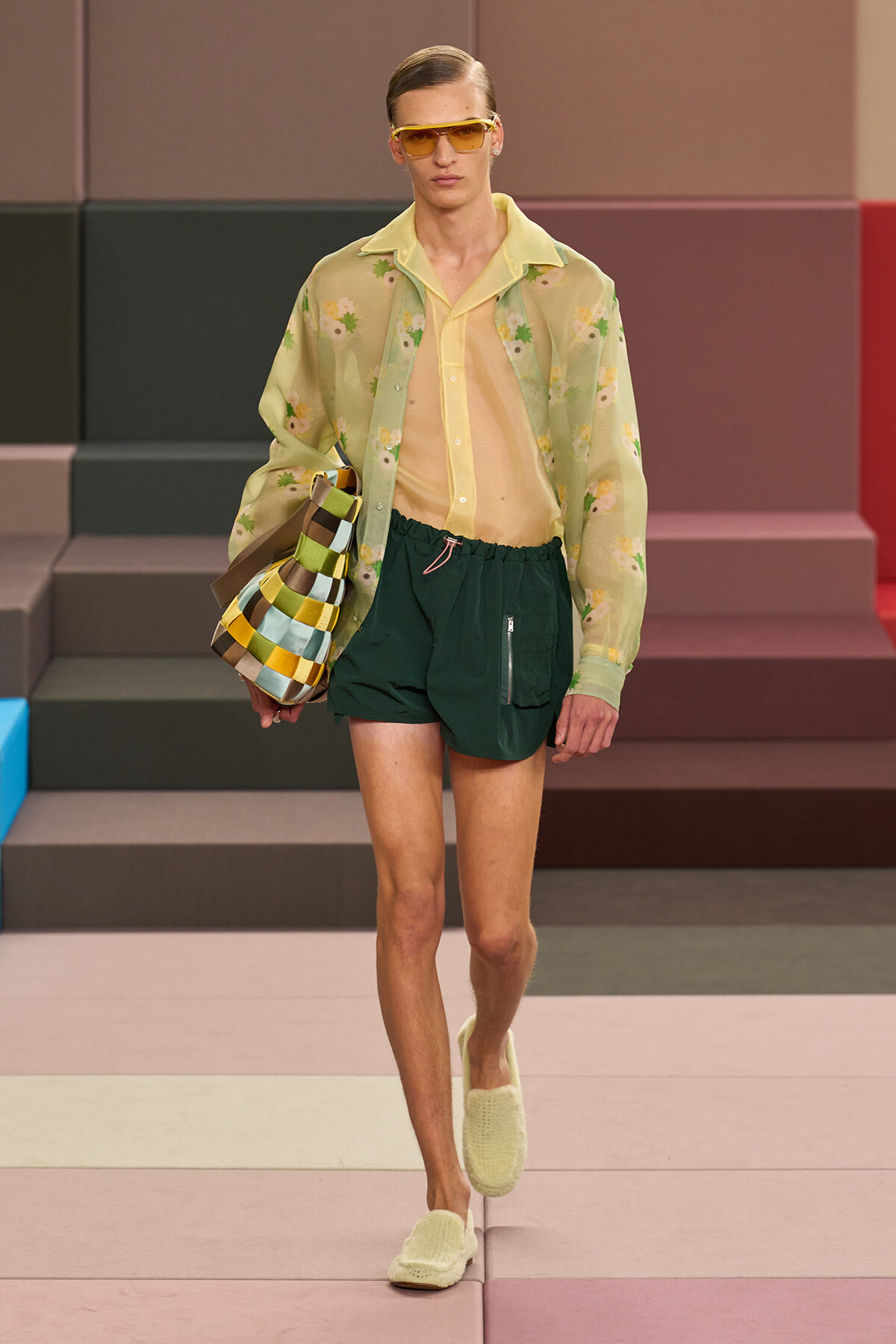 Male model walks a runway in a sheer yellow floral shirt over a pale top, dark green shorts, yellow slides, orange sunglasses, and a multicolored striped bag.