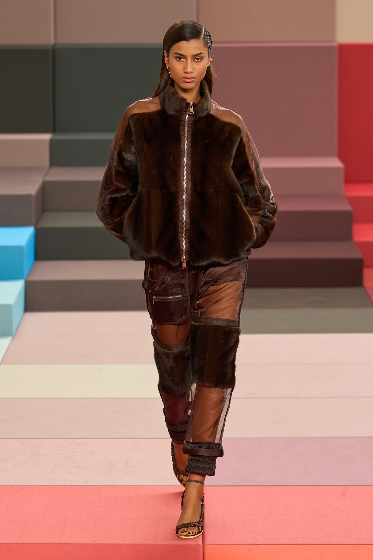 Model walks the runway in a dark brown fur zip-front jacket and patchwork pants with sheer panels against a colorful stepped backdrop.