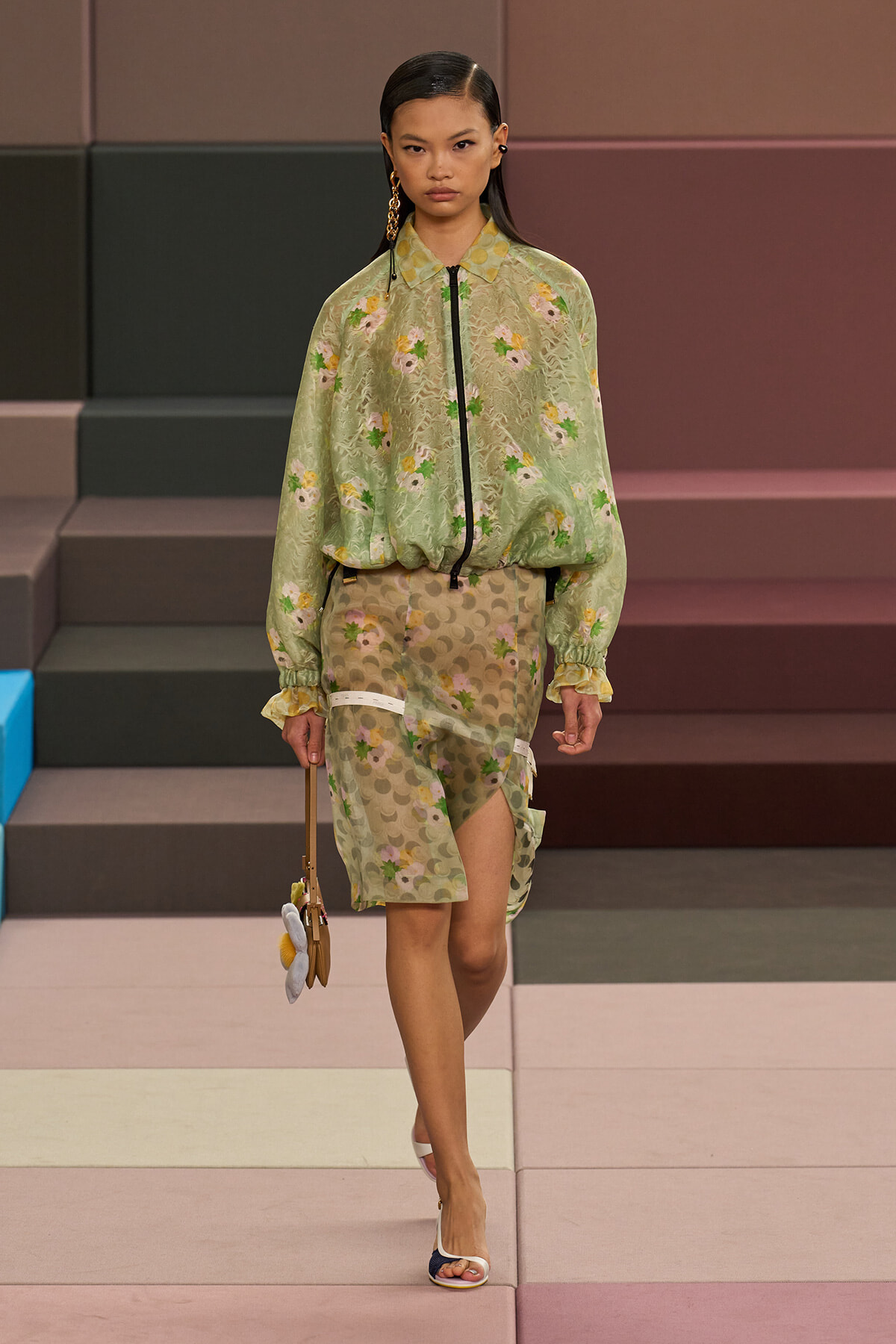 Model walking a runway in a sheer sage-green floral blouse with a black zipper paired with a beige patterned midi skirt featuring a side slit and white bands, holding a small handbag with fuzzy pom-poms and white sandals.