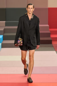 Male model walks a fashion runway in a black blazer with matching shorts, carrying a small multicolored quilted bag, wearing woven black loafers on a pastel geometric catwalk.
