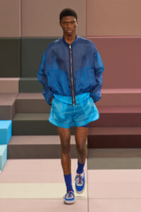 Male model walks a fashion runway in blue athleisure: navy bomber jacket, bright blue shorts, matching socks, and blue-white sneakers.