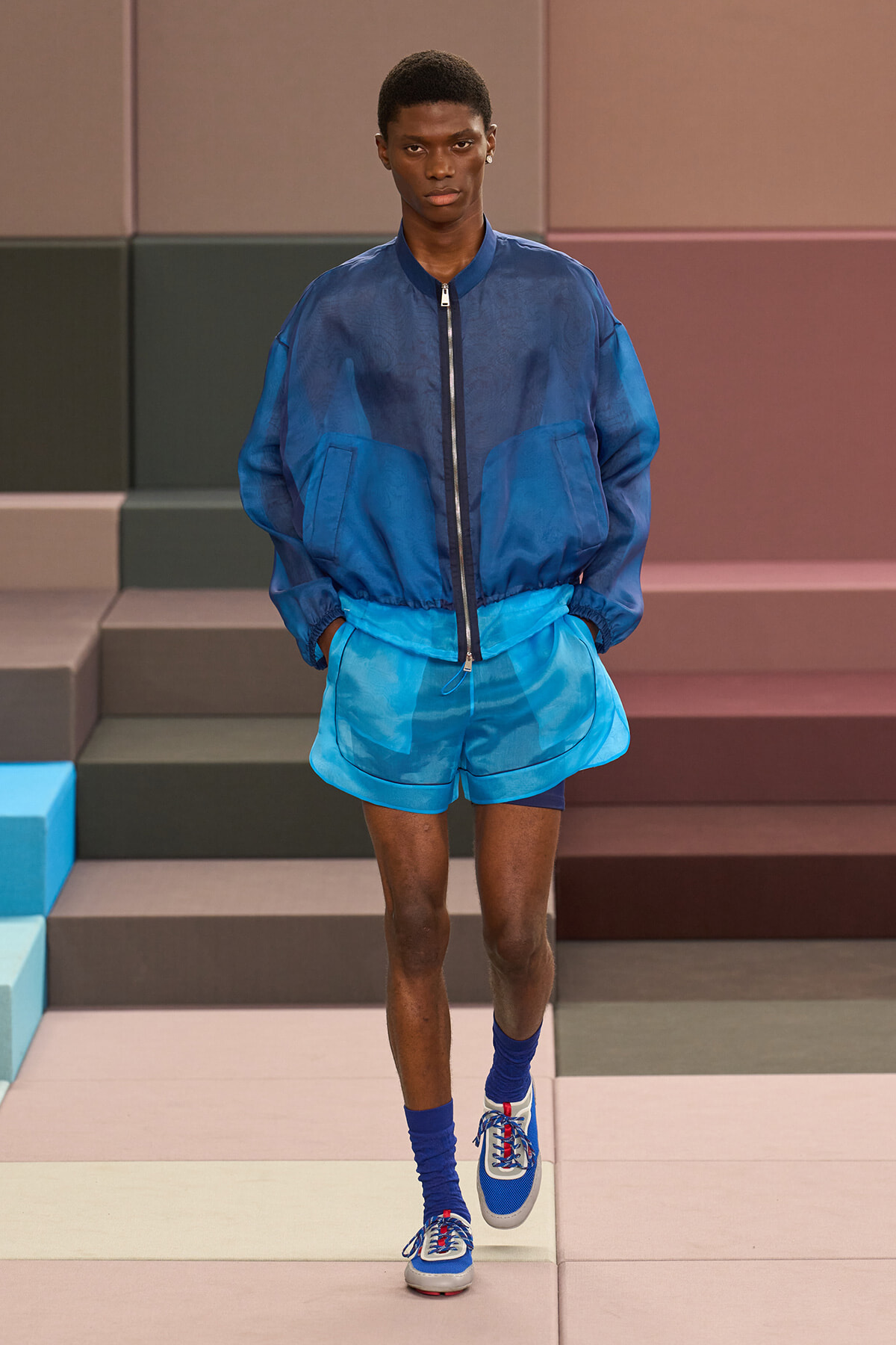 Male model walks a fashion runway in blue athleisure: navy bomber jacket, bright blue shorts, matching socks, and blue-white sneakers.