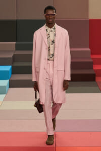 Male model in a pale pink suit with matching trousers, floral shirt, and sunglasses walks a colorful runway during a fashion show.