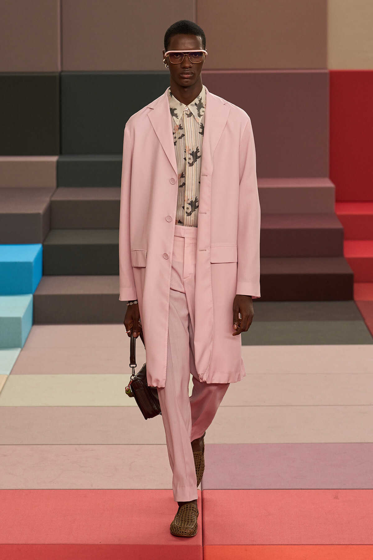 Male model in a pale pink suit with matching trousers, floral shirt, and sunglasses walks a colorful runway during a fashion show.