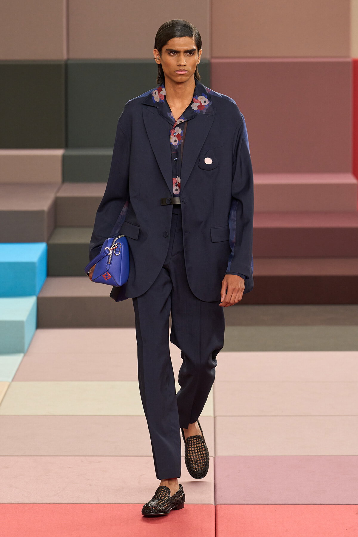 Male model walks a pastel-tiled runway in a navy suit with a floral shirt and a blue bag slung under his arm.