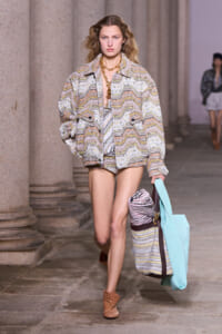 Model walks a runway in a pastel zigzag coat, brown boots, and gold necklace, carrying turquoise tote and a striped rolled bag.