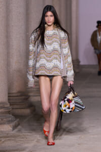 Model walks the runway in a chevron-knit tunic with a matching coat, carrying a multicolored woven handbag.