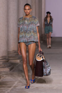 Model walking a runway in a sequined multicolor top with matching shorts, carrying a large patterned tote and a burgundy bag while wearing blue open-toe heels.