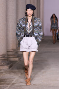 Model on a runway wearing a multicolored zigzag knit jacket, white cuffed shorts, a black belt, brown ankle boots, and a navy beret.