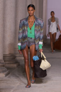 Model walking a runway in a multicolor metallic blazer, green top, matching shorts, and chunky gold necklace while carrying a tote bag and a knit item wrapped in a dark bag.