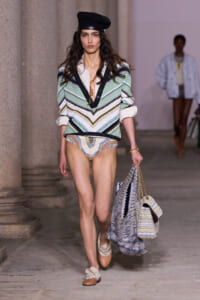 Model walking a runway in a green-and-cream chevron knit sweater, white shirt cuffs, patterned bikini bottoms, tan shoes, a black beret, and carrying a large woven tote bag.