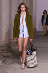 Model walking a runway in an olive green utility jacket over a light blue striped shirt, carrying a large woven tote bag.