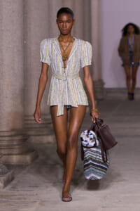 Model walking a runway in a pastel striped belted mini dress with puff sleeves, layered gold necklace, burgundy tote, and patterned bag under her arm.