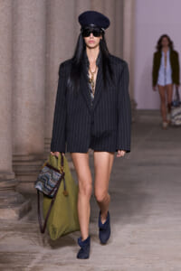 Runway look: model wears oversized pinstripe blazer as a short dress, navy hat, dark sunglasses, gold necklace, blue ankle boots, and carries a large olive-green bag with a patterned panel.