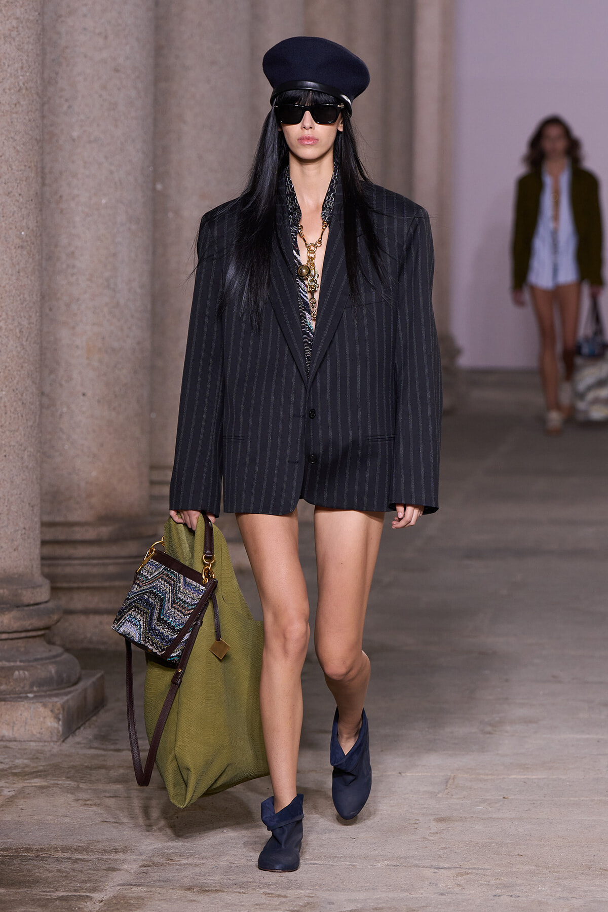 Runway look: model wears oversized pinstripe blazer as a short dress, navy hat, dark sunglasses, gold necklace, blue ankle boots, and carries a large olive-green bag with a patterned panel.