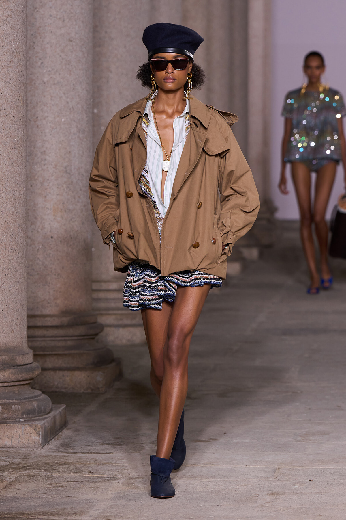 Runway model wearing a tan oversized trench over a striped blouse and textured mini skirt, with a navy cap and sunglasses.