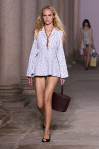 Model walks the runway in a pale blue textured sleeveless coat, black sandals, and a dark maroon bucket bag; layered gold necklace visible.