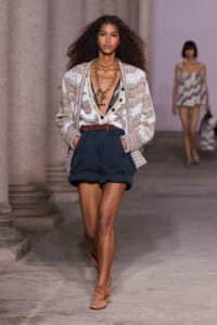 Model walking a runway in a patterned cardigan over black high-waisted shorts with a brown belt and gold necklace; tan flats, loose curly hair.