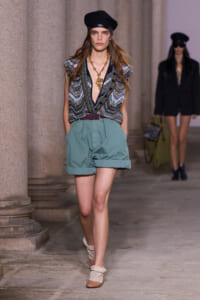 Model walking the runway in teal rolled-cuff shorts, a deep V zigzag-knit top, black beret and chunky gold necklace.