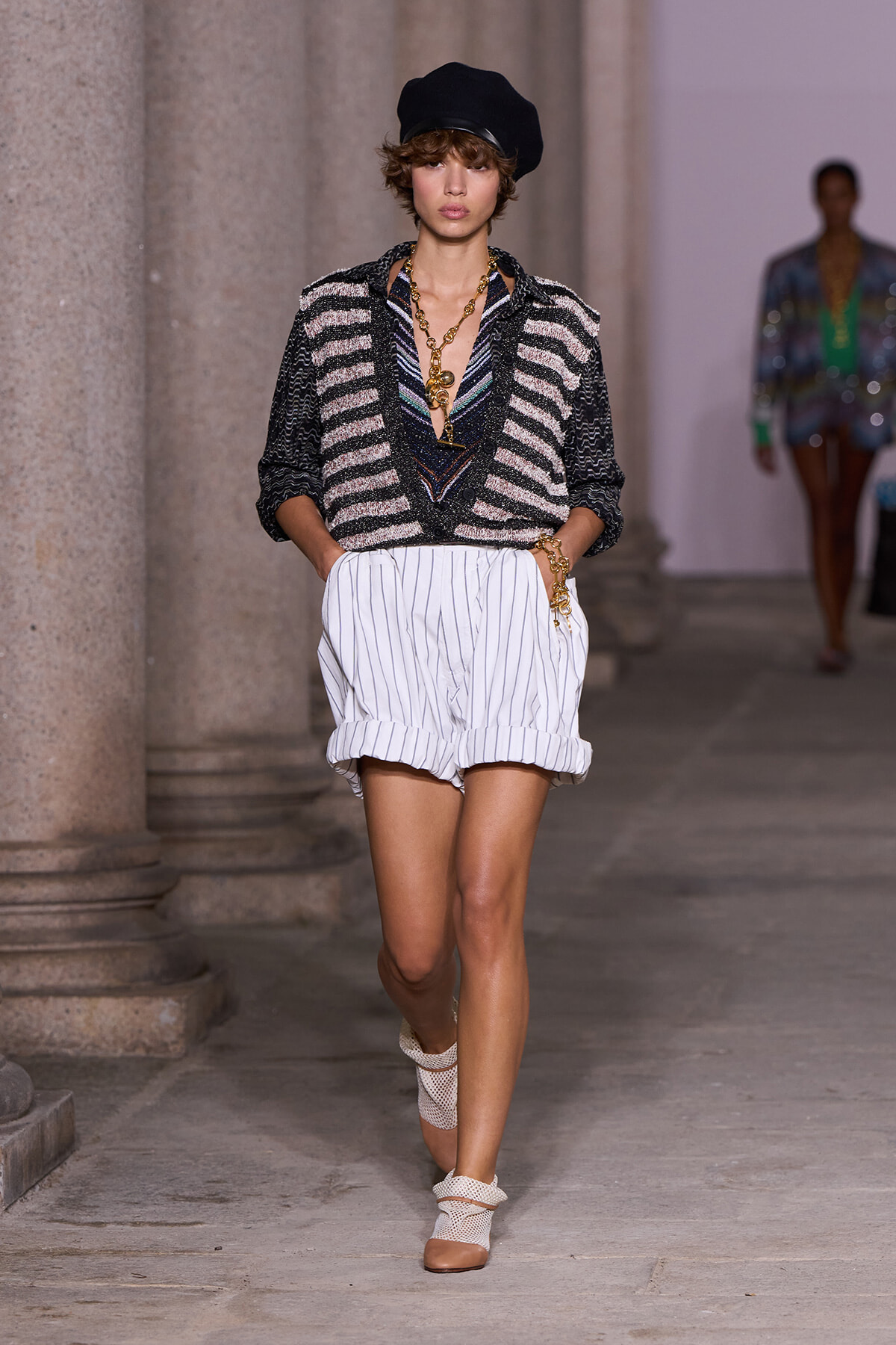 Model walks a runway wearing a black beret, striped black-and-white blouse, white pinstripe shorts, and chunky gold jewelry.