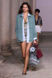 Model walks the runway in a teal utility jacket over a blue-and-white striped shirt, with a multicolor woven tote and burgundy handbag.