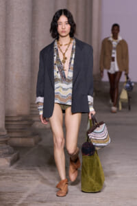 Model walking a runway in an oversized dark blazer over a patterned V-neck top, bikini bottoms, long gold necklace, brown ankle boots, and carrying a large green bag with a striped purse and rolled textiles.
