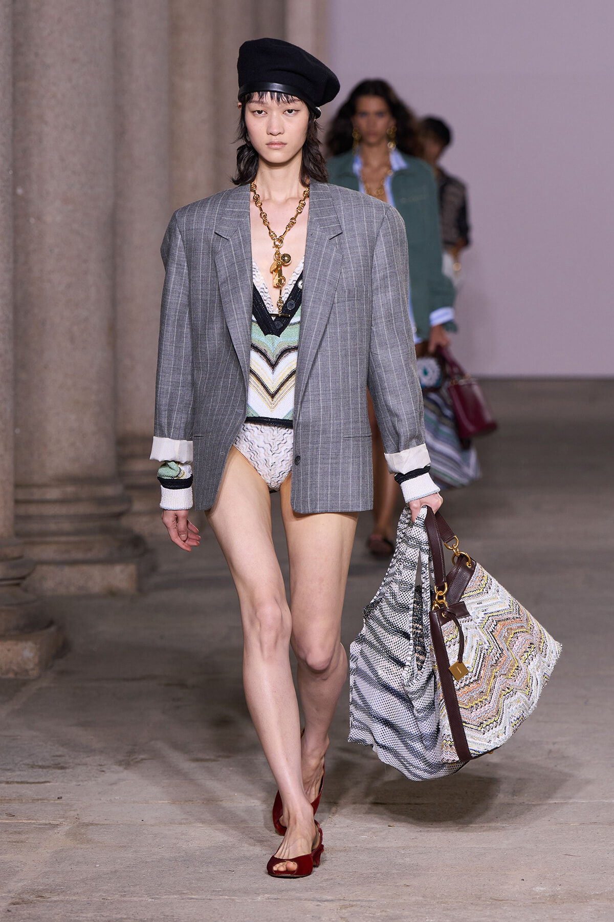 Model on runway wearing a gray pinstripe blazer over a colorful patterned bodysuit, black beret, chunky gold necklace, and burgundy flats, carrying a large zigzag-print tote bag.