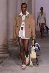Model walking runway in tan field jacket over striped knit top and white shorts, carrying a large beige bag with a patterned scarf.