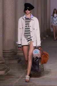Model walking a runway in a beige utility jacket over a black-and-white striped dress, accessorized with a black hat, oversized sunglasses, and gold jewelry, carrying a large brown suede bag with a rolled knit blanket and a patterned tote.