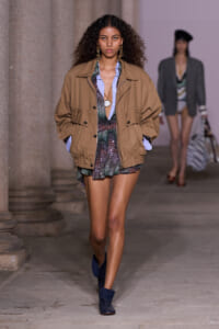 Model walking runway in a tan oversized jacket over a shimmery multicolor dress and blue shirt, wearing blue ankle boots and gold earrings