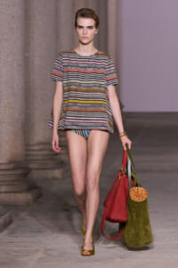 Model walking a runway in a multicolor striped short-sleeve top over swim bottoms, carrying red and olive-green tote bags with a rolled orange item inside.