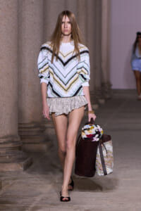 Model walking the runway in a white chevron-striped sweater and beige ruffled shorts, carrying a burgundy tote and a round knit bag with a colorful pattern.