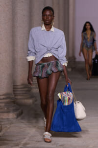Model walking a runway beside stone columns in a gray knotted sweater over a striped collared shirt, green ruffled shorts, and cream sandals, carrying a large blue bag with striped towel and a cream knit bag
