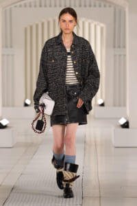 Model walking a runway in a textured black tweed jacket over a striped top, black mini skirt, tall layered boots, and a white handbag with a red chain.