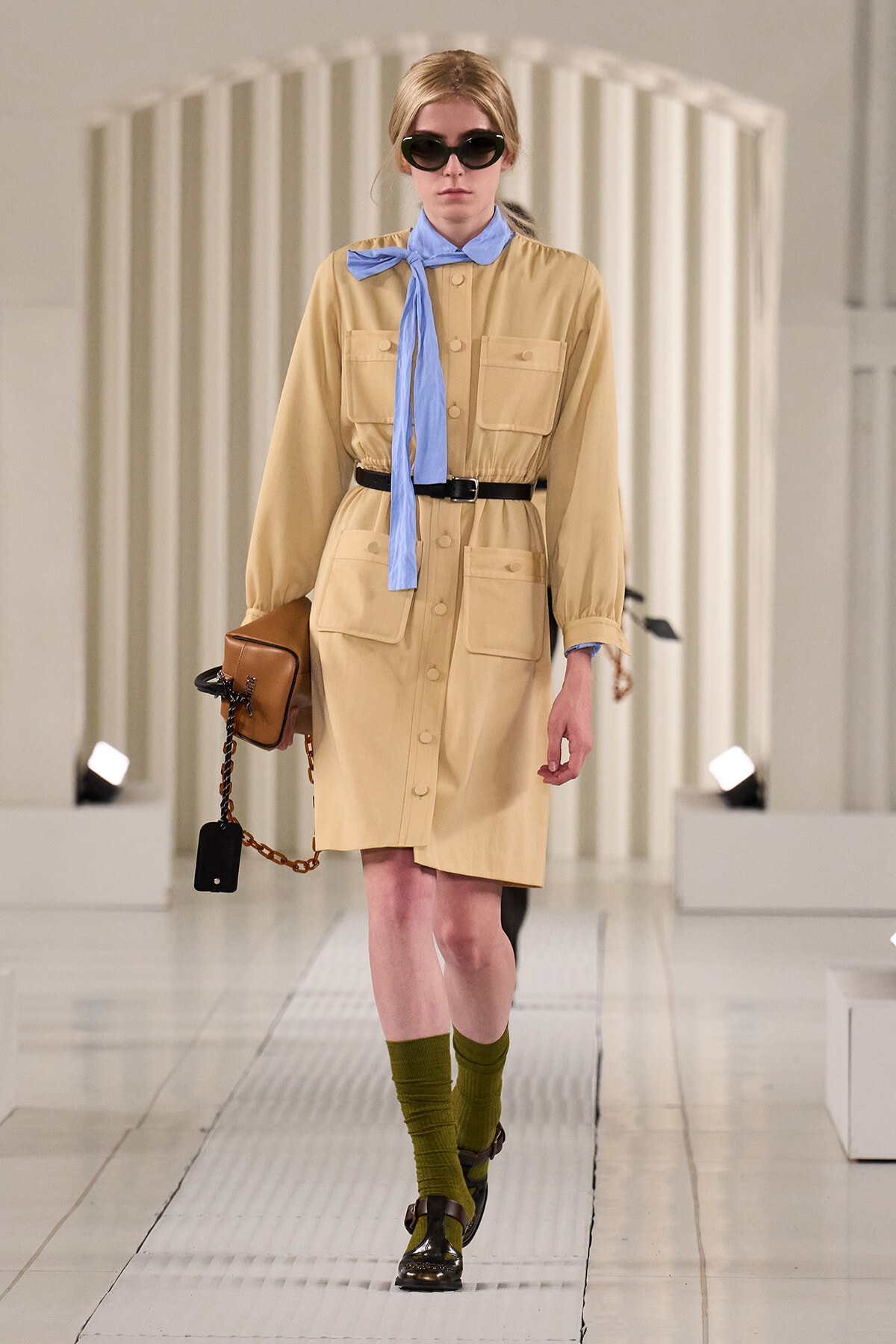 Model walks the runway in a tan utility shirt-dress with large pockets, a blue neck scarf, black belt, green socks, brown shoes, sunglasses, and a brown handbag with a chain.