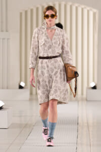 Model walking a fashion runway in a pale floral shirt-dress with a brown belt, scarf, and oversized sunglasses, carrying a tan handbag with a chain strap.
