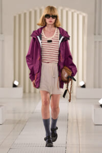Model on a runway wearing a purple jacket over a striped cream top, beige drawstring shorts, gray socks, black sandals, oversized sunglasses, and a brown bag with a chain.