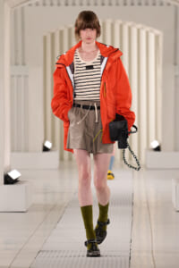 Model on a runway wearing an orange rain jacket over a striped cream top and beige shorts, with olive socks and black sandals, carrying a black bag with a chain.
