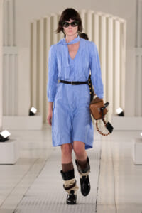 Female model walking a fashion runway in a light blue belted shirt dress with a black belt, oversized sunglasses, and two-tone boots, carrying a brown handbag with a chain.