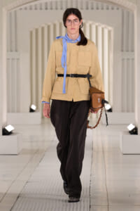 Model walking runway in a beige utility jacket with large pockets, blue neck scarf, black belt, and black trousers, carrying a brown crossbody bag with a chain strap.
