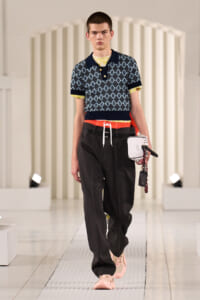 Male model walking a fashion show in a patterned polo shirt, dark pants, pink sneakers, and a red drawstring belt, while holding a small white bag with a chain.