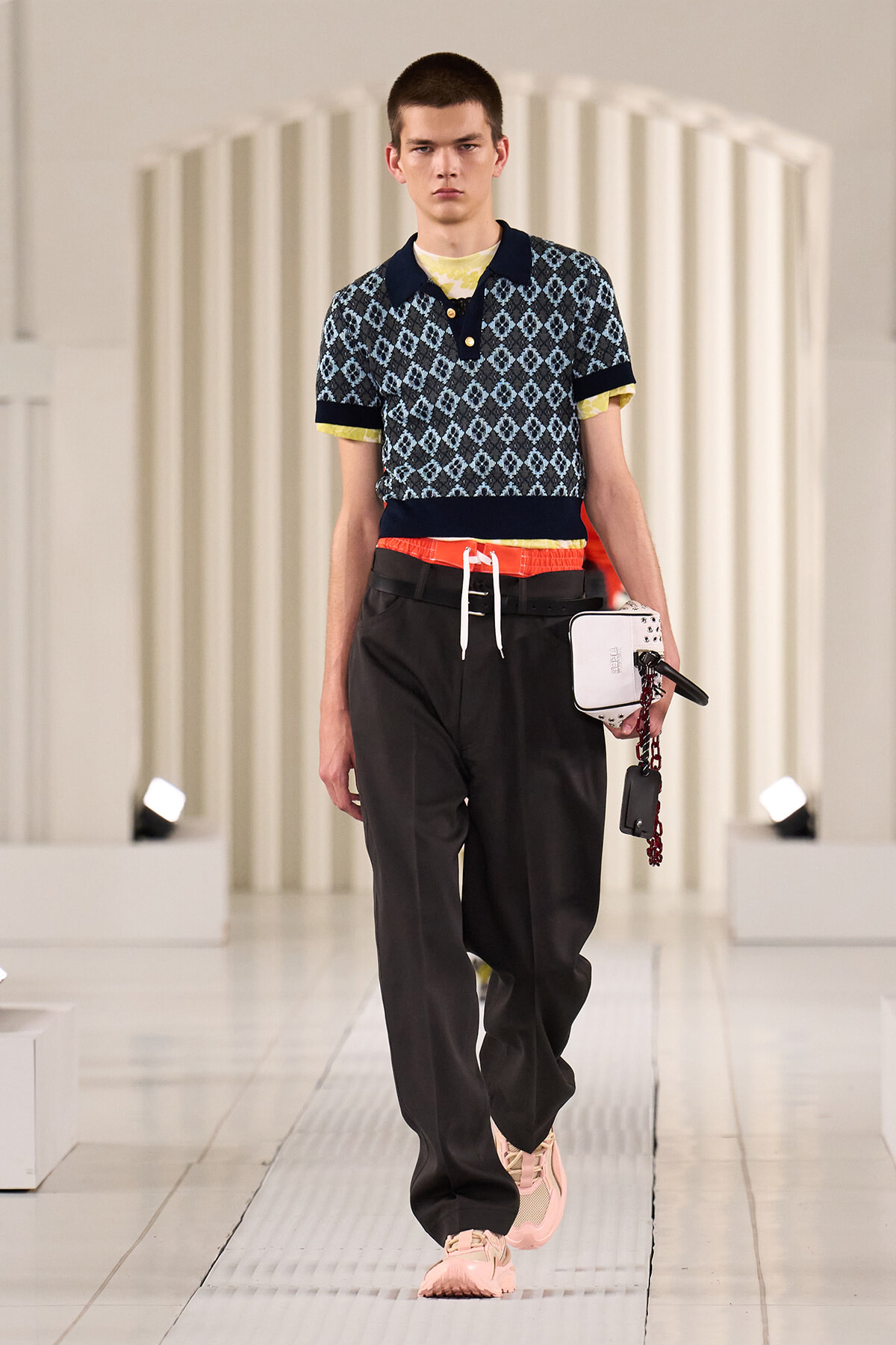 Male model walking a fashion show in a patterned polo shirt, dark pants, pink sneakers, and a red drawstring belt, while holding a small white bag with a chain.
