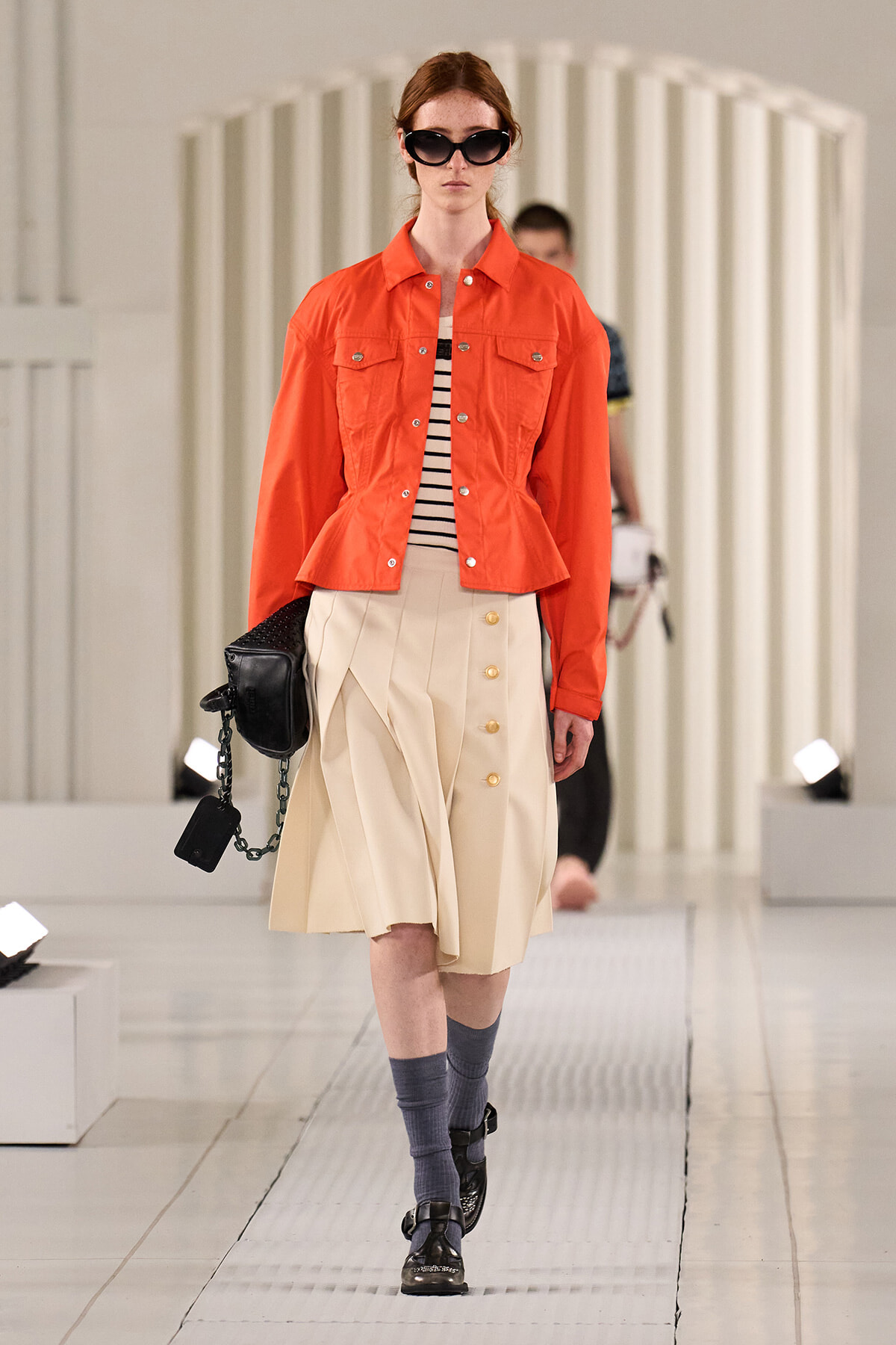 Model walking on a runway in an orange cropped jacket, striped top, beige button-front skirt, dark sunglasses, and black accessories.